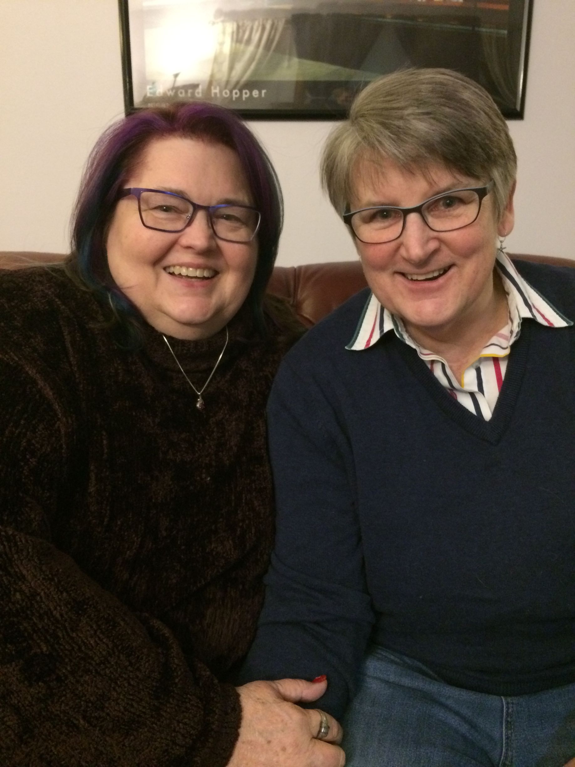 Two people smiling side-by-side, one with purple hair in a dark sweater and one in a navy sweater, holding hands.