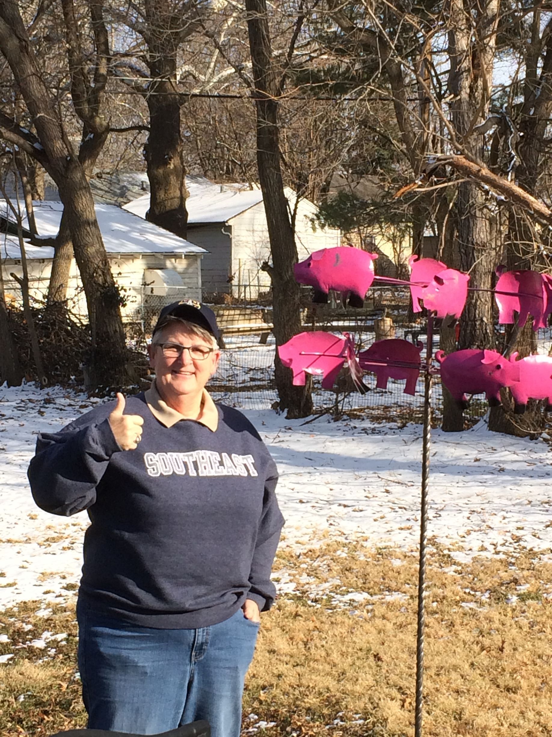 Person in a navy sweatshirt giving a thumbs up in a snowy yard featuring several hanging, bright pink pig-shaped signs.