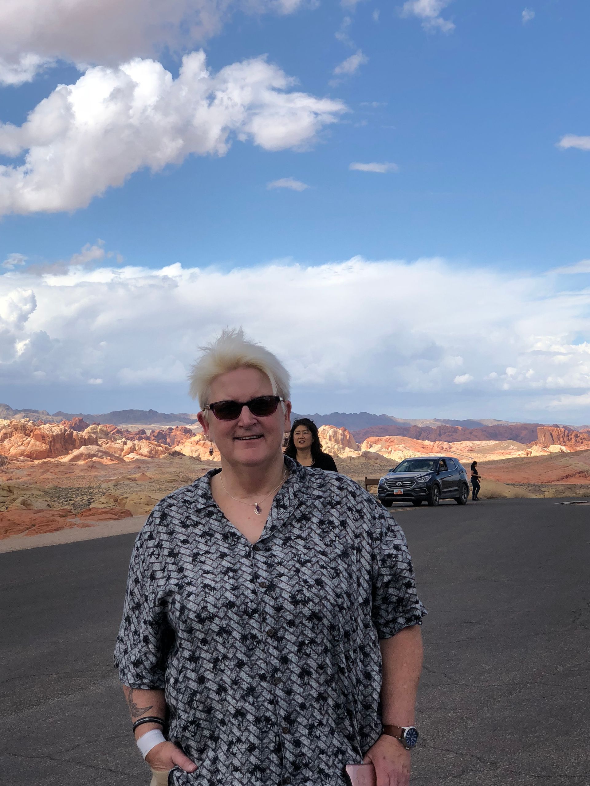 A person with short light hair and sunglasses smiles while standing on a desert road with red rock formations in the back.
