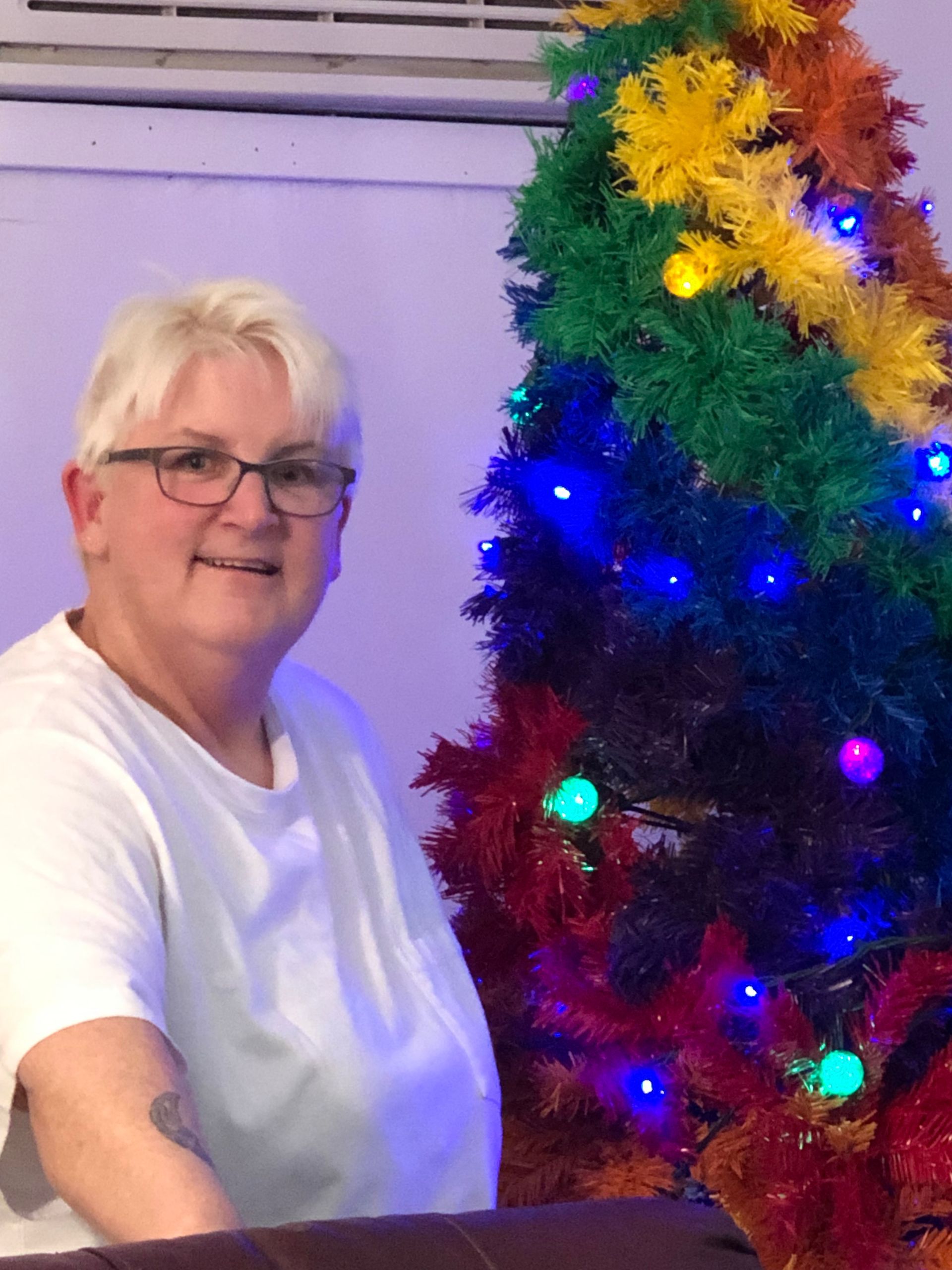 A person in a white shirt smiles next to a colorful, lit-up rainbow Christmas tree in an indoor setting.