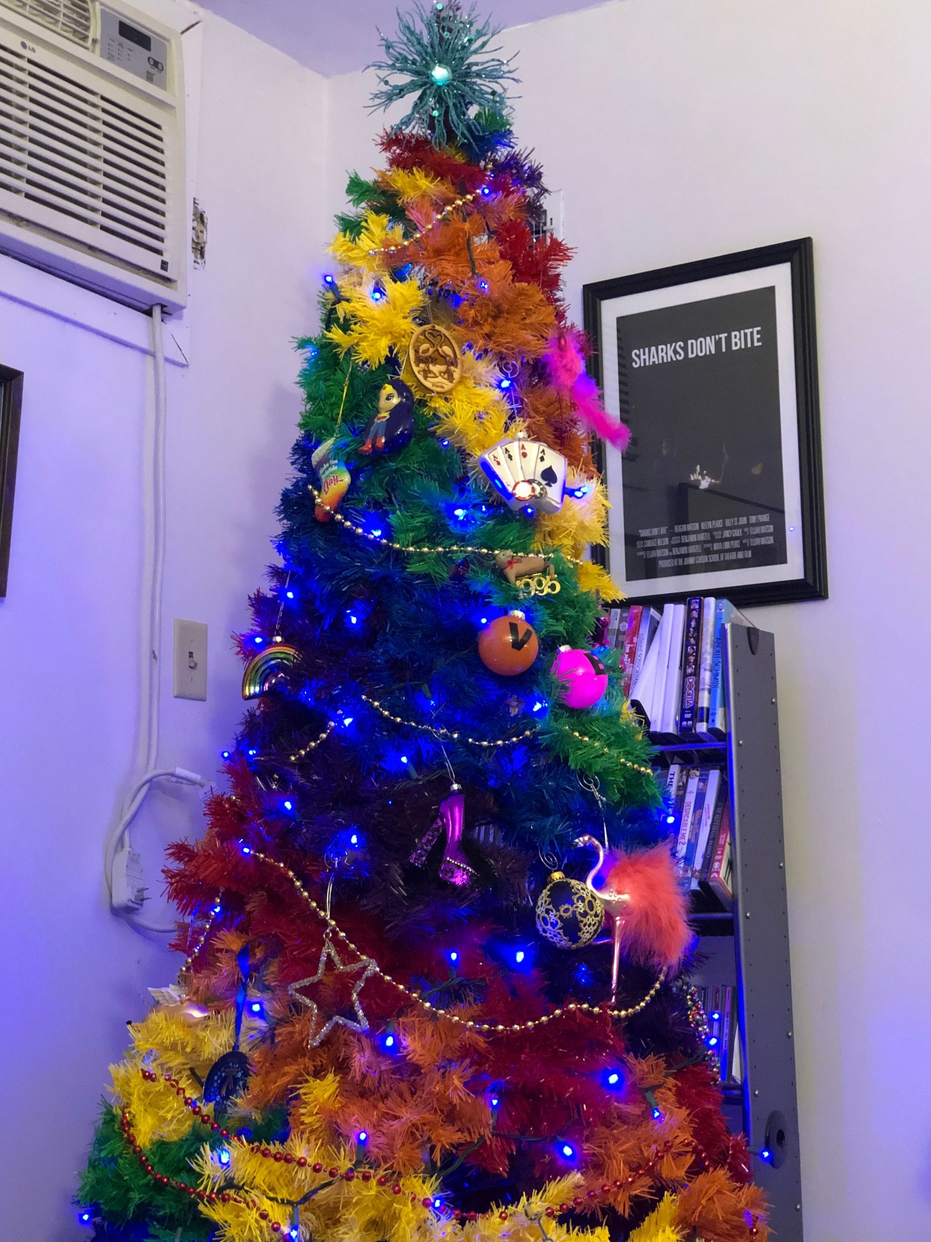 A rainbow-colored Christmas tree decorated with blue string lights, standing in a corner next to a framed sign.
