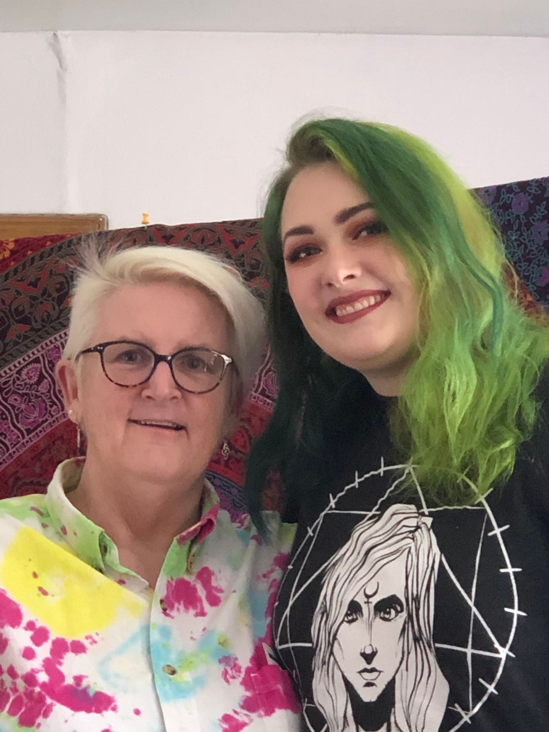Two people smiling together against a patterned tapestry; one wears a tie-dye shirt, the other has vibrant green hair.