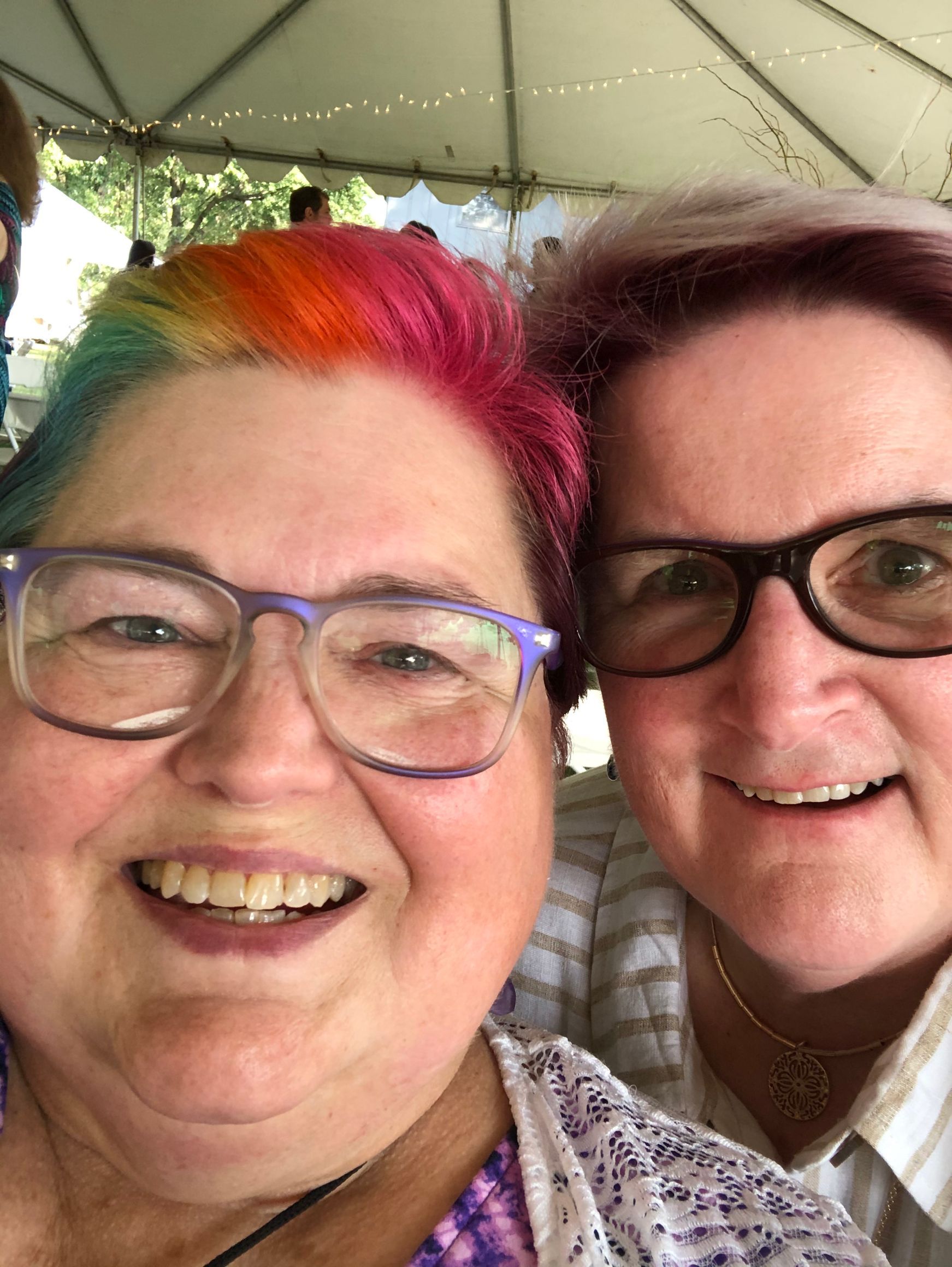 Two smiling people pose for a selfie under a tent canopy. One has rainbow-colored hair and the other has short mauve hair.