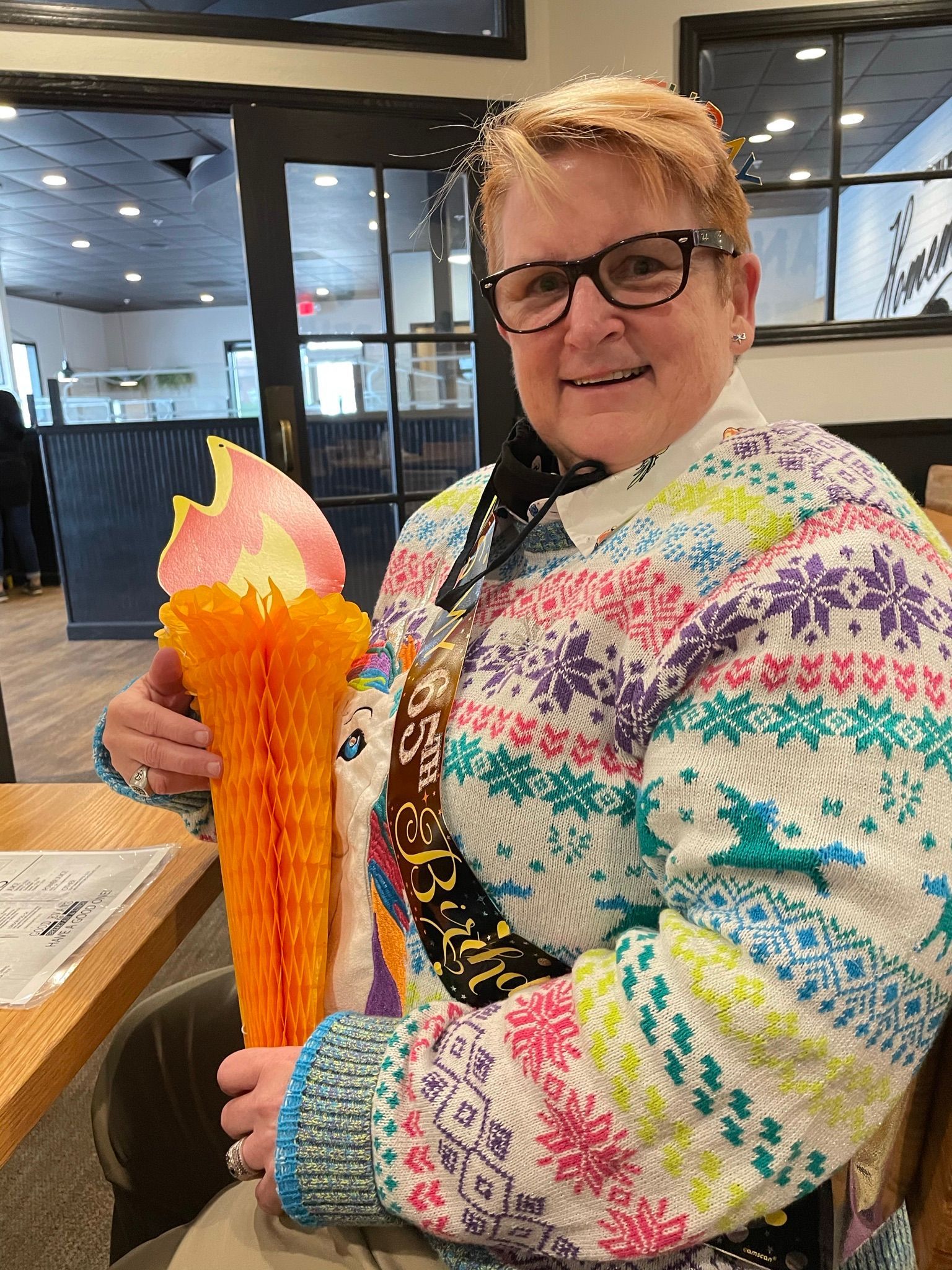 A person wearing glasses and a colorful patterned sweater holds a decorative paper torch at a table.