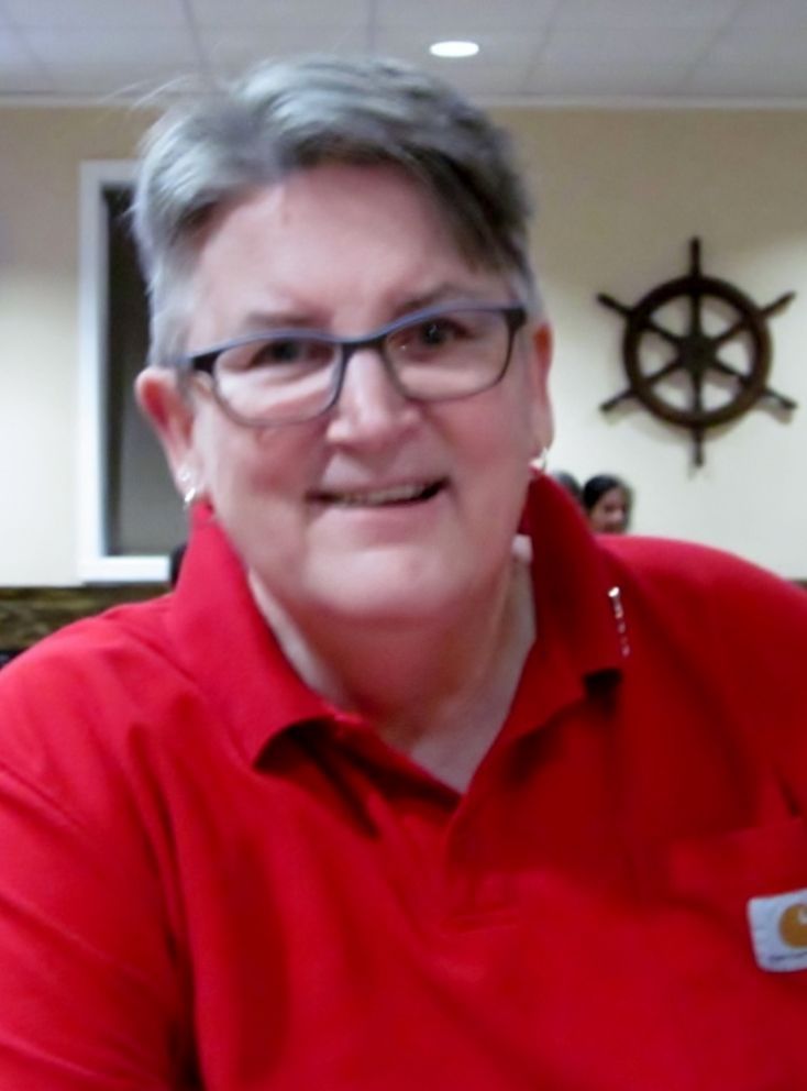 A smiling person with short grey hair and glasses wears a red polo shirt, positioned in front of a nautical wall decor.