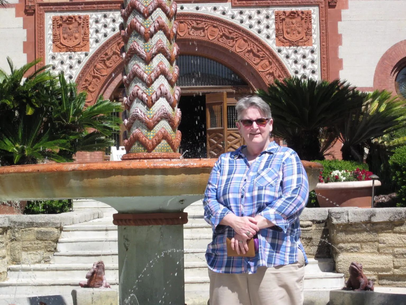 A person in a blue plaid shirt stands in front of a stone fountain with a decorative pillar, near a building entrance.
