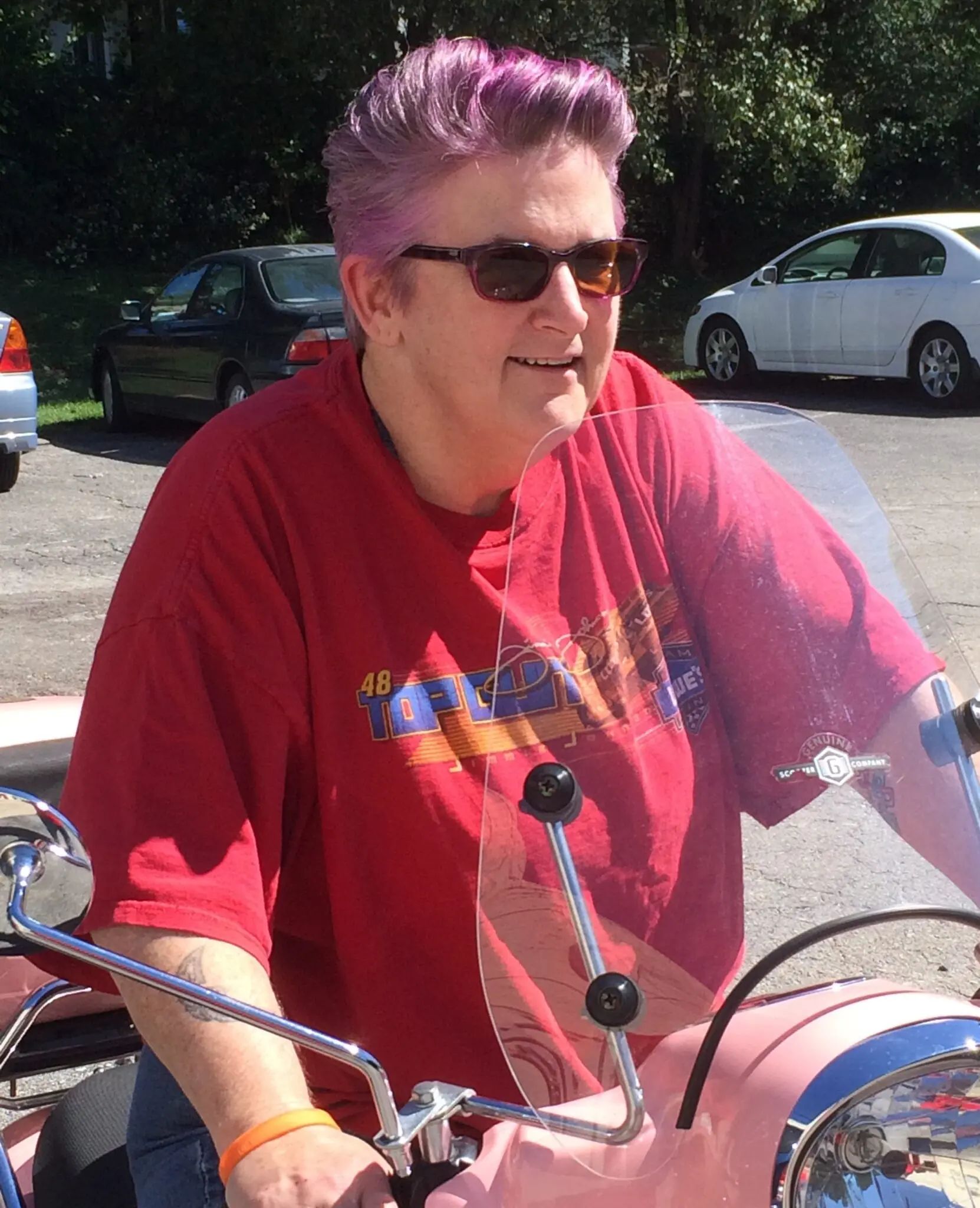 A person with short purple hair and sunglasses smiles while sitting on a pink motorcycle in an outdoor parking lot.