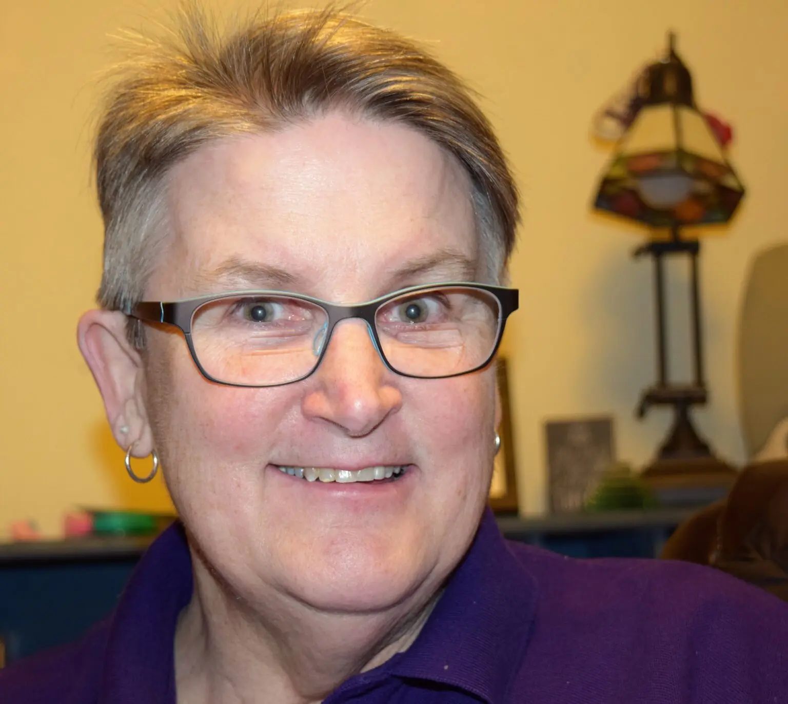 A person with short, light hair wearing dark-rimmed glasses and a purple shirt, smiling in an indoor setting.