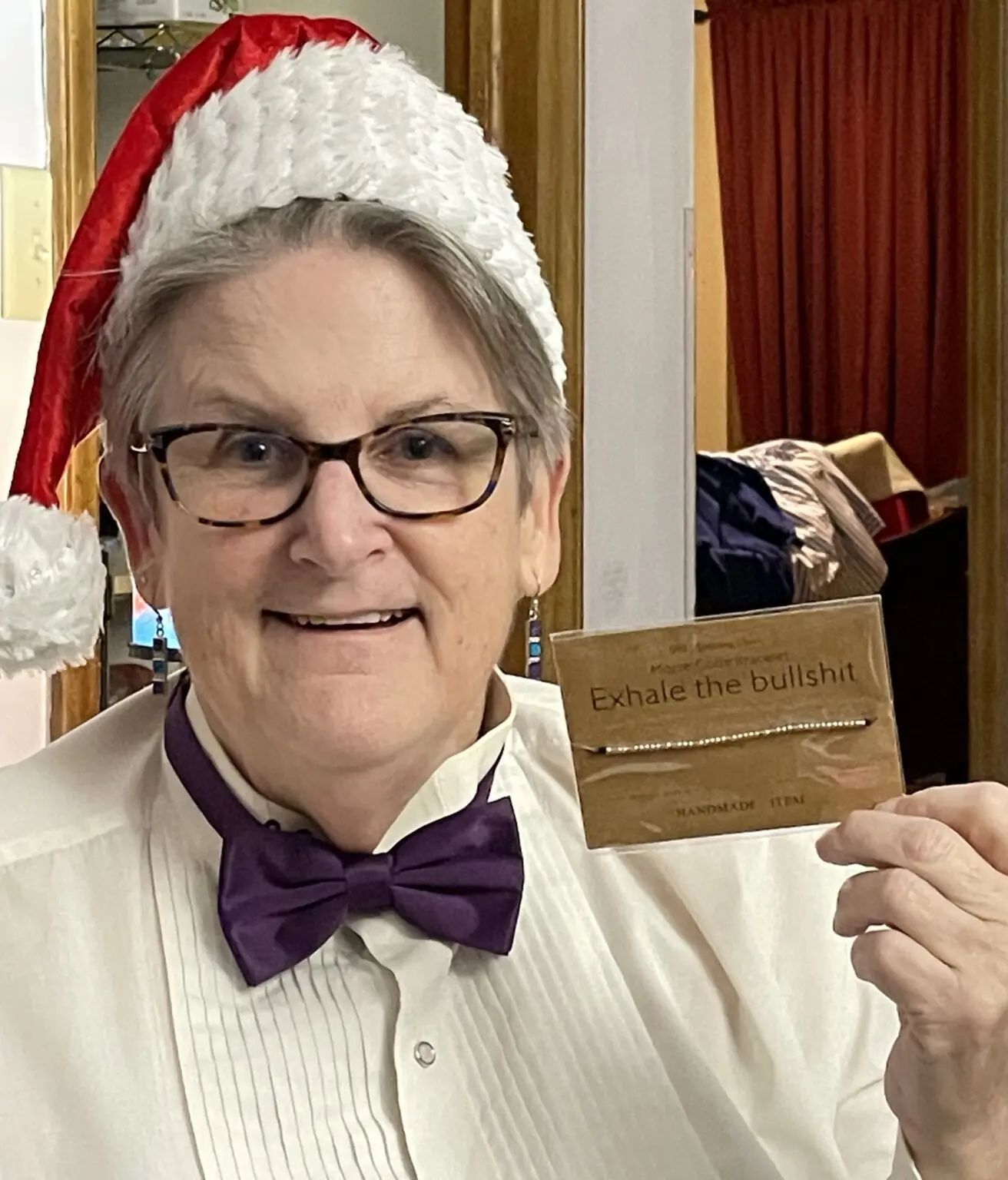 A person in a Santa hat and purple bow tie holds a small card that reads 