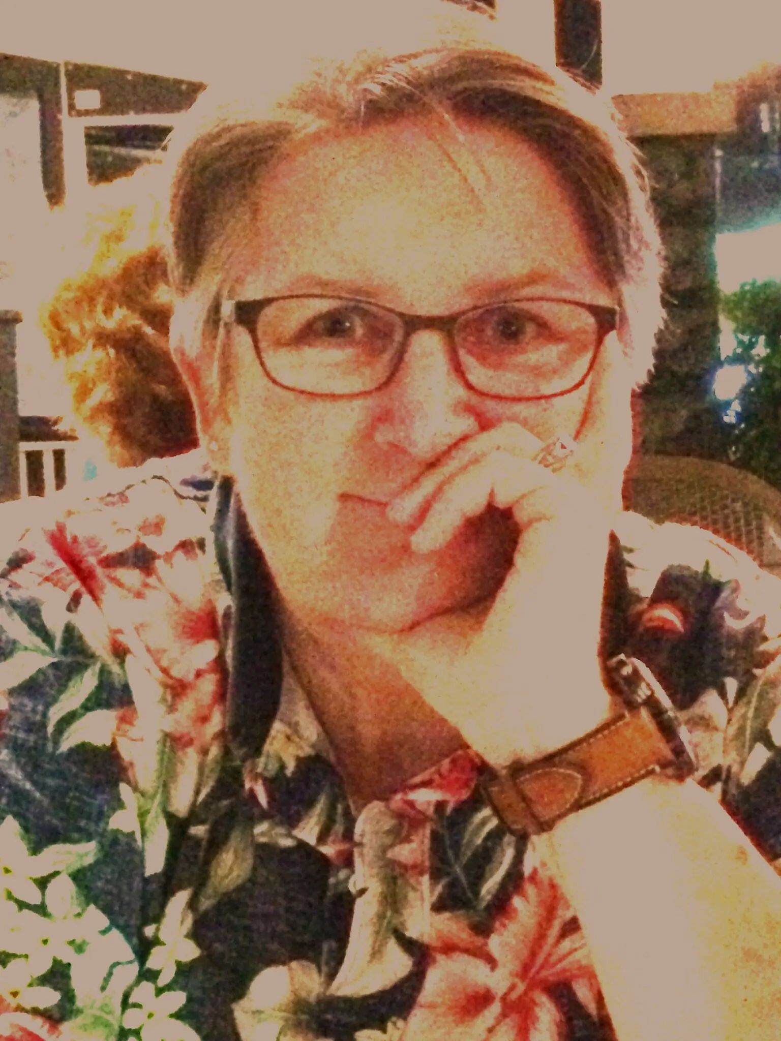 A person wearing glasses and a floral shirt rests their chin on their hand while looking toward the camera.