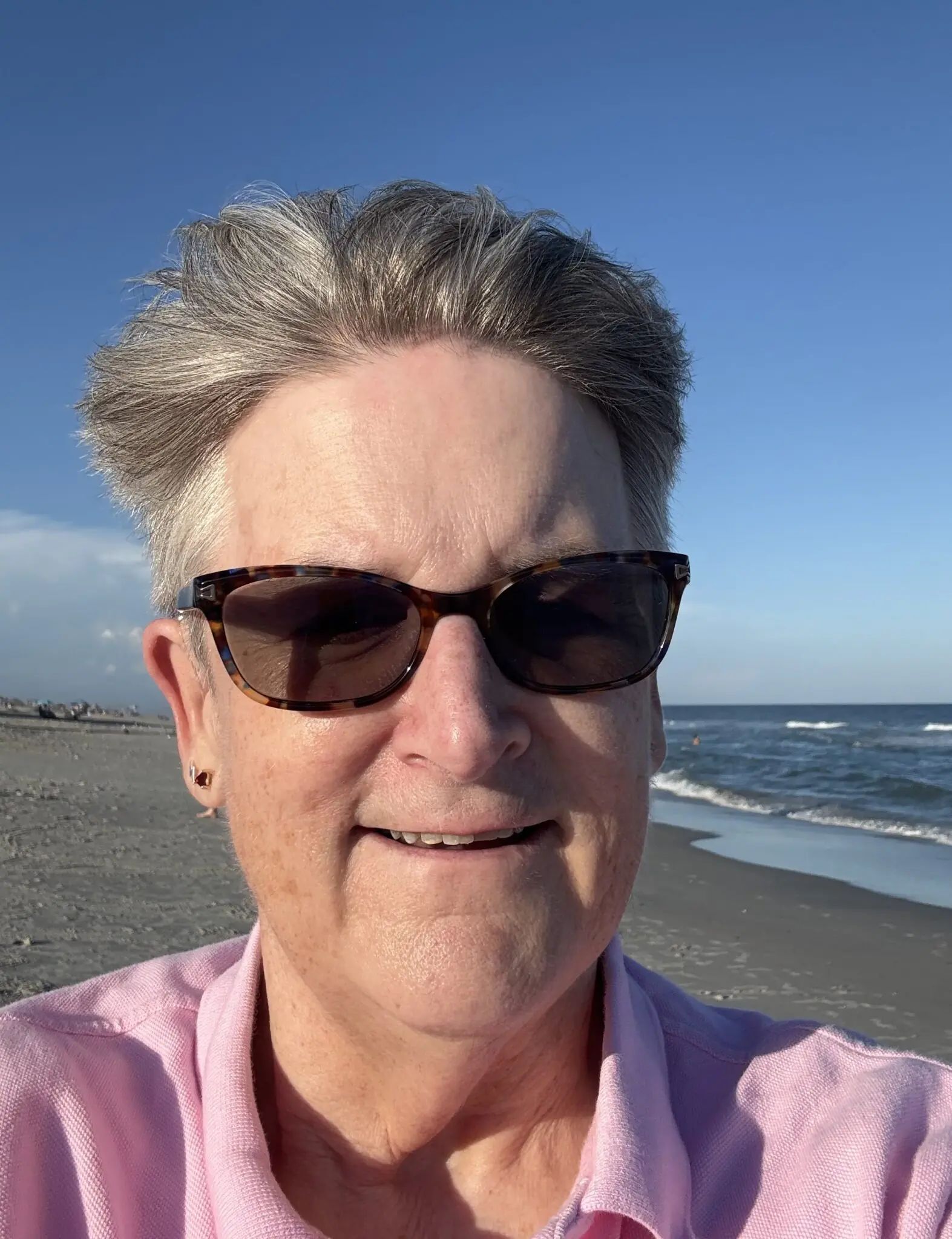 A person wearing sunglasses and a pink shirt smiles while posing on a beach with the ocean and blue sky in the background.