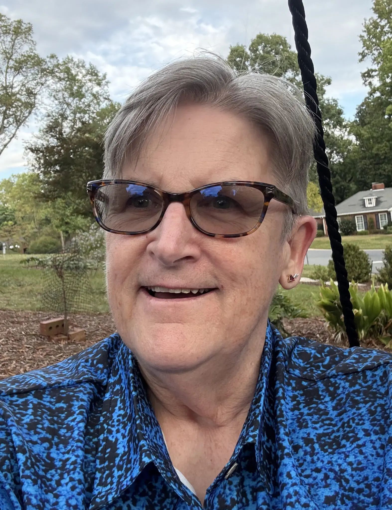 A person with short grey hair and patterned glasses smiling in front of a house and trees.