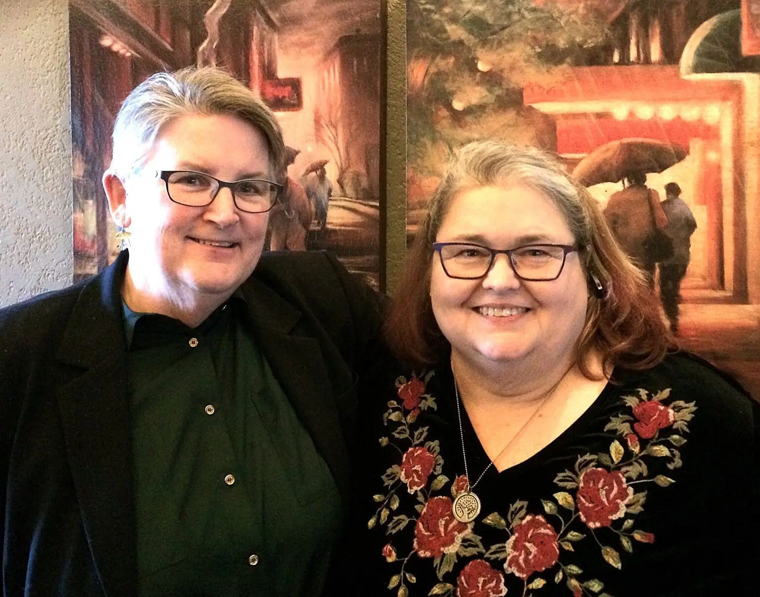 Two smiling people stand side-by-side in front of two paintings of a street scene, both wearing glasses.