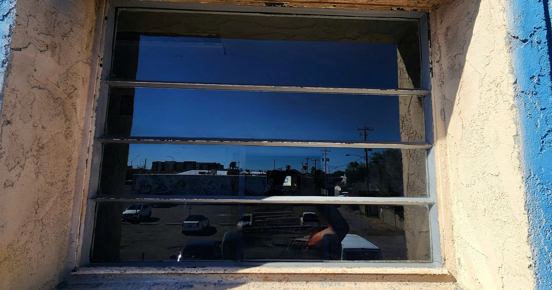 Window reflecting blue sky, street scene, and person inside a building.