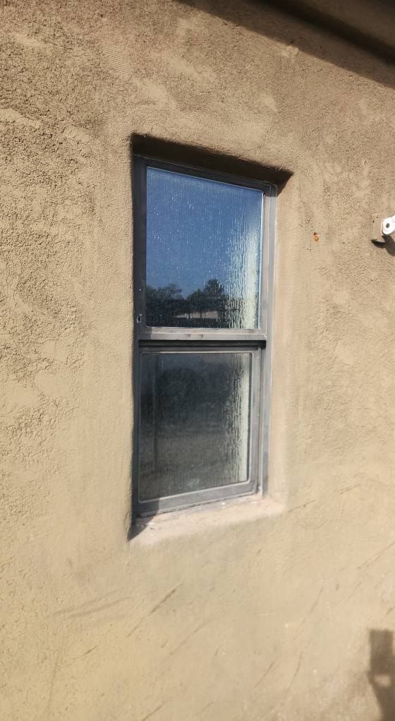 Vertical window on a stucco wall. The glass reflects sky and trees. The wall is light brown.