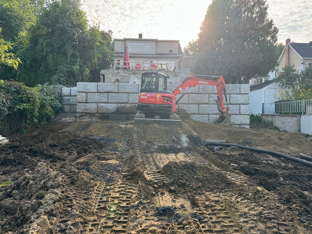 Une excavatrice rouge roule sur un chemin de terre devant une maison.