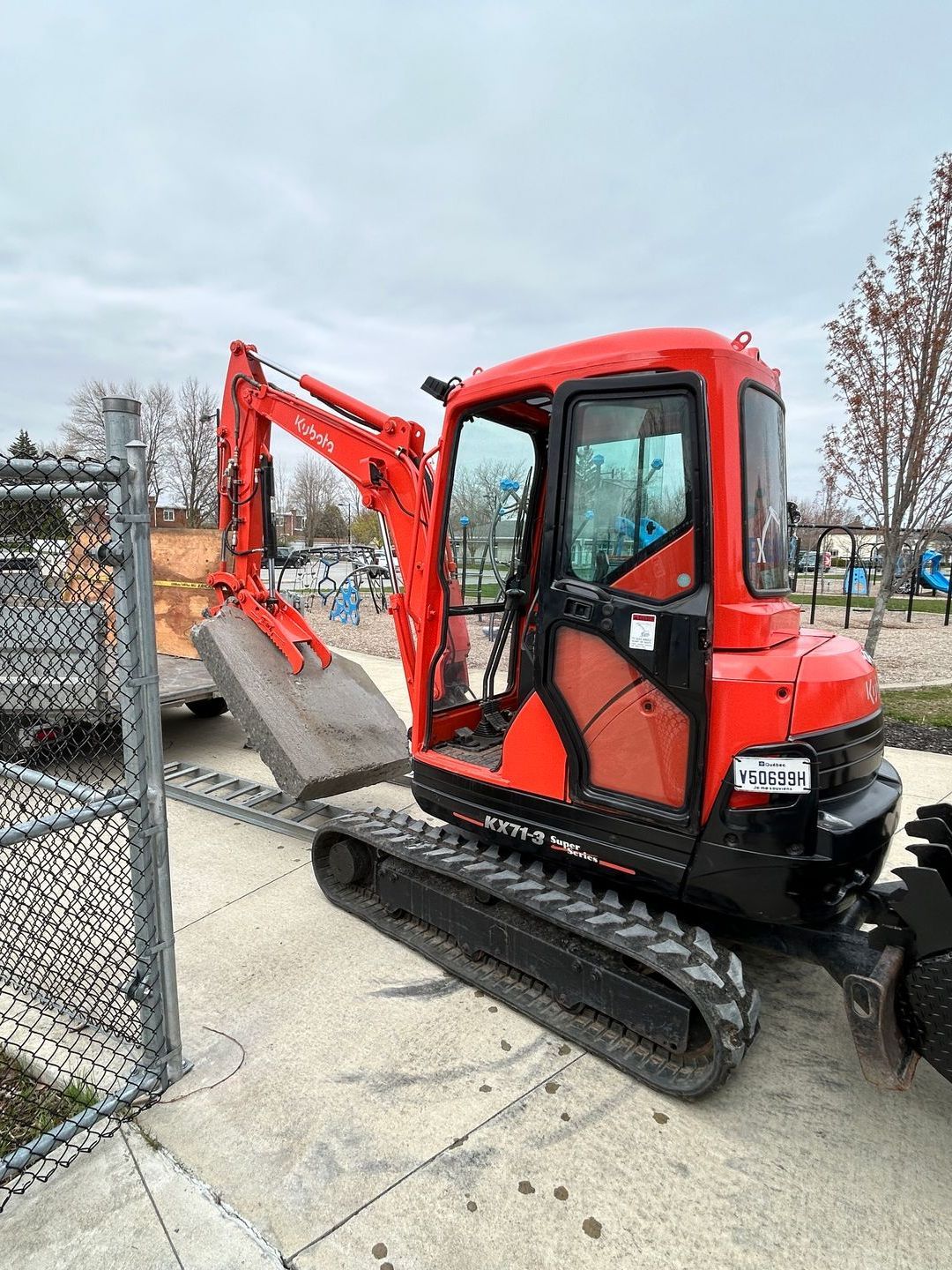 Une petite excavatrice rouge est garée sur le trottoir à côté d'une clôture grillagée.