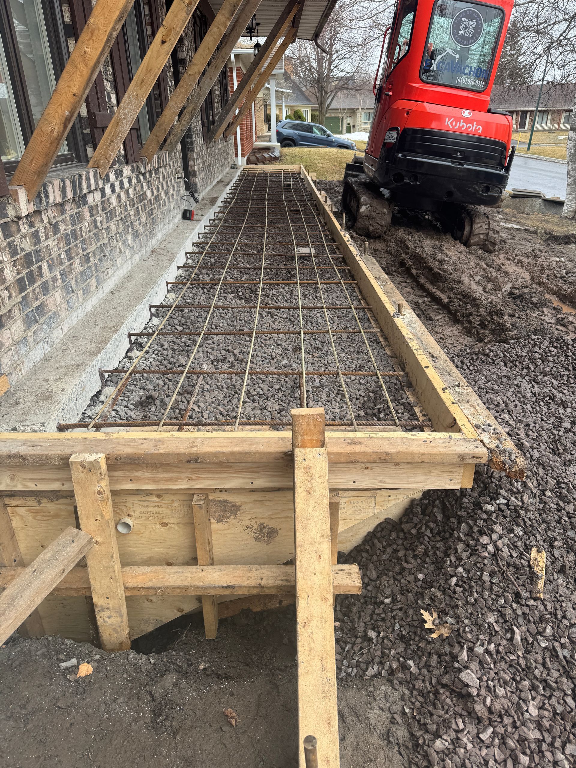 Une excavatrice rouge roule sur un chemin de terre à côté d'une fondation en béton.
