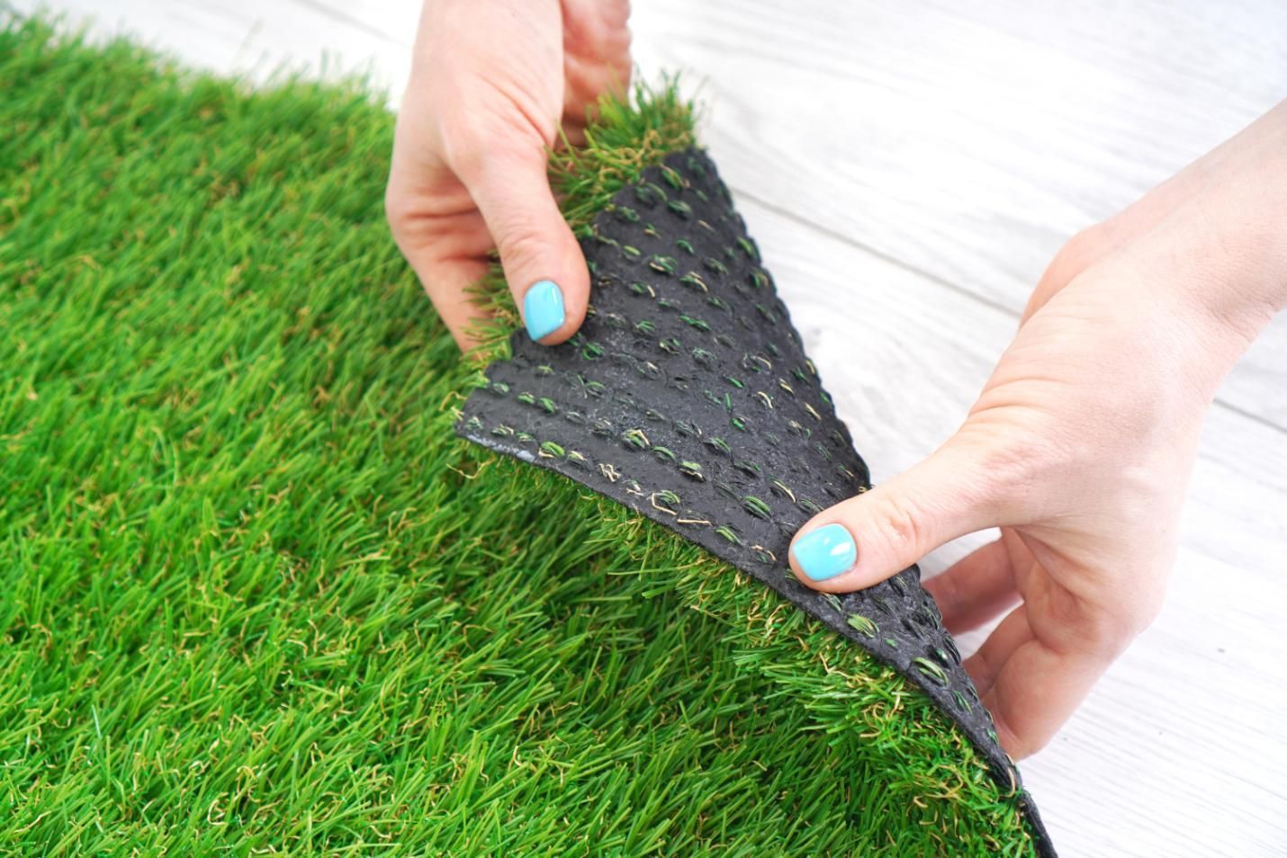Hands holding up a corner of artificial turf, revealing its black backing and green blades.