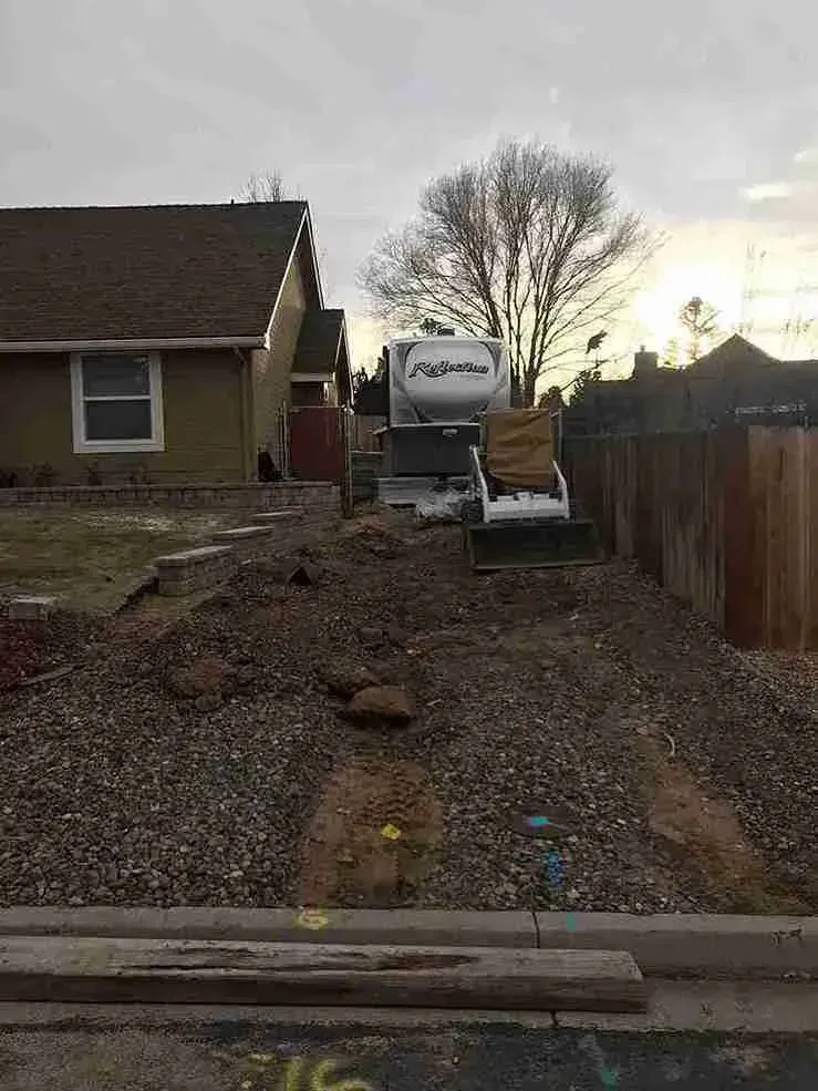 Pile of gravel in front yard with RV, near house and wooden fence.
