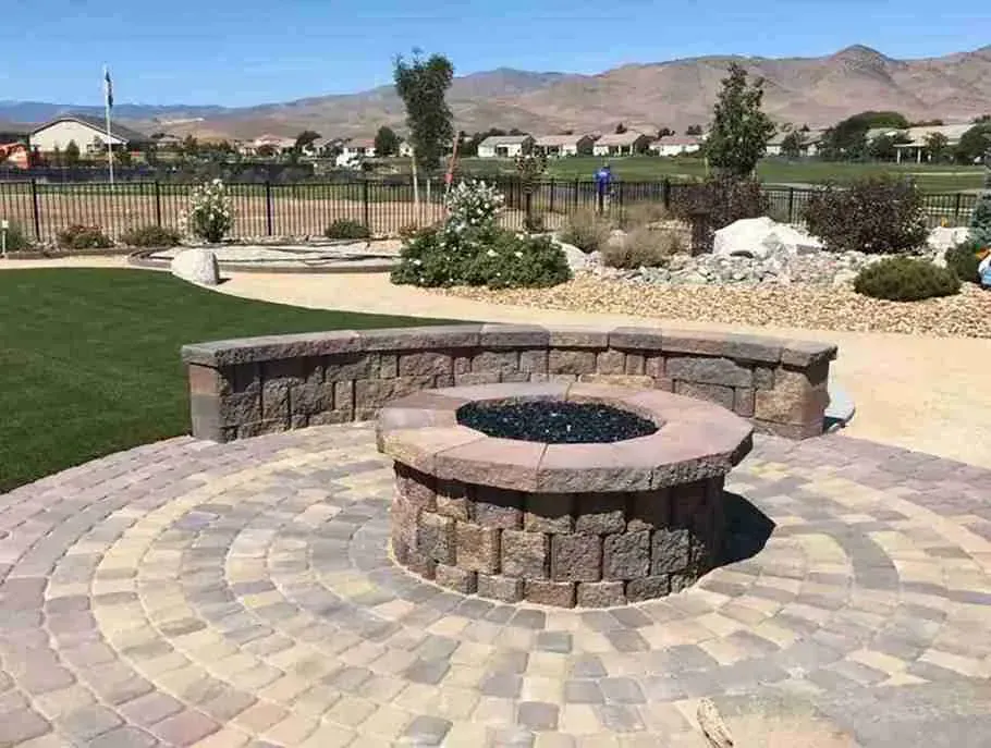 Fire pit on a circular stone patio with retaining wall, green grass, and mountains in the background.