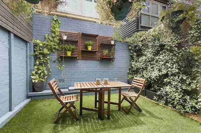 Wooden table and chairs on artificial turf in a small, enclosed backyard with plants and a decorative wall.