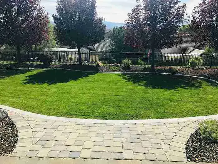Brick pathway leads to a green lawn with trees and a fenced yard under a blue sky.