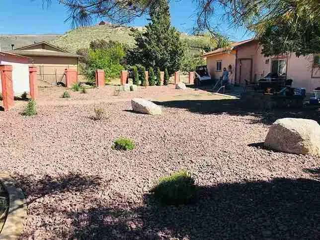 Yard with reddish gravel, large rocks, small bushes, and two buildings.
