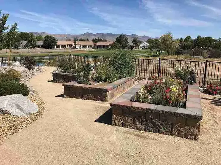 Raised garden beds with various plants, set on tan gravel with a lake and houses in the background.