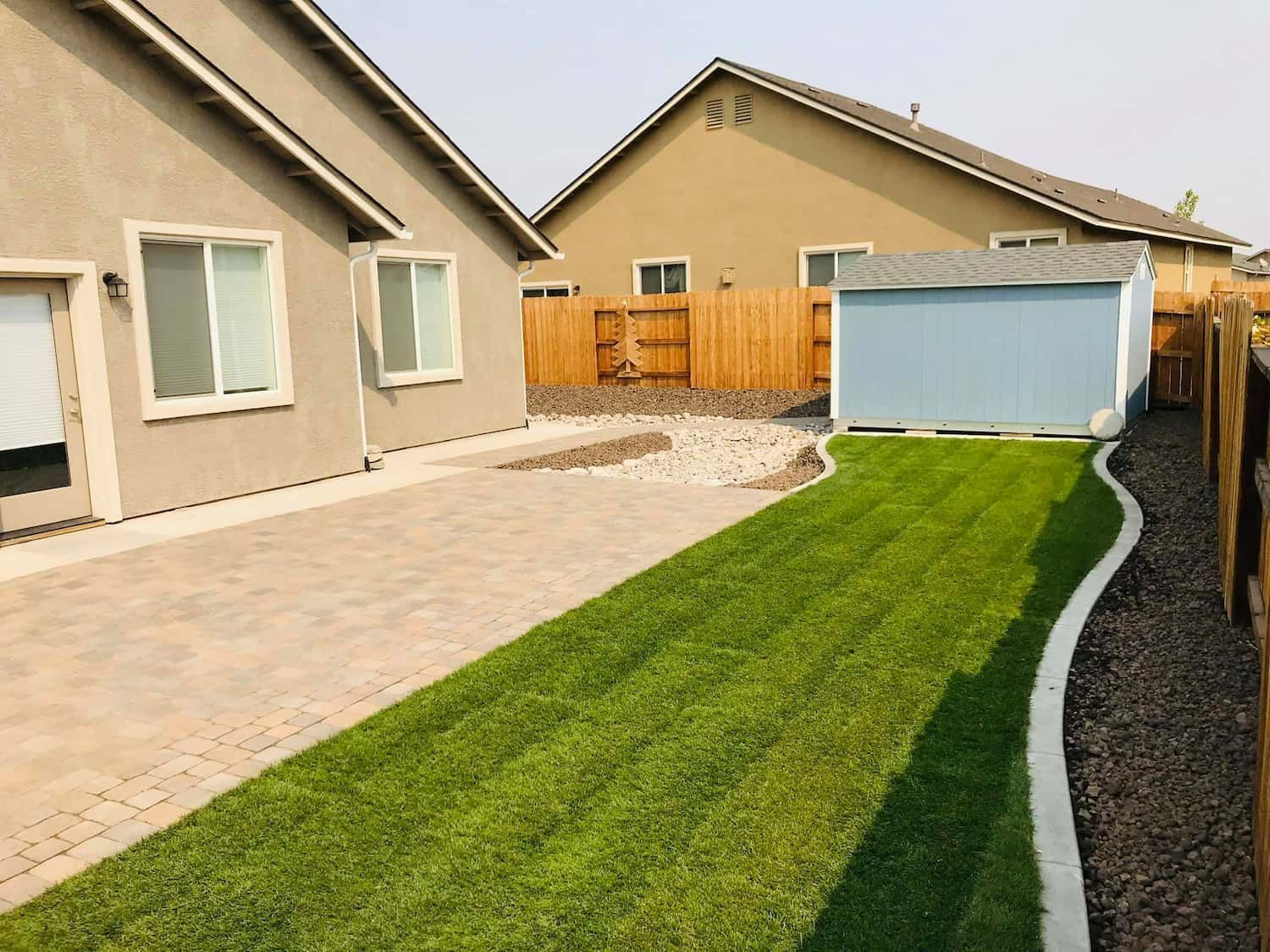Backyard with a light blue shed, green grass, a paved patio, and a brown wooden fence.