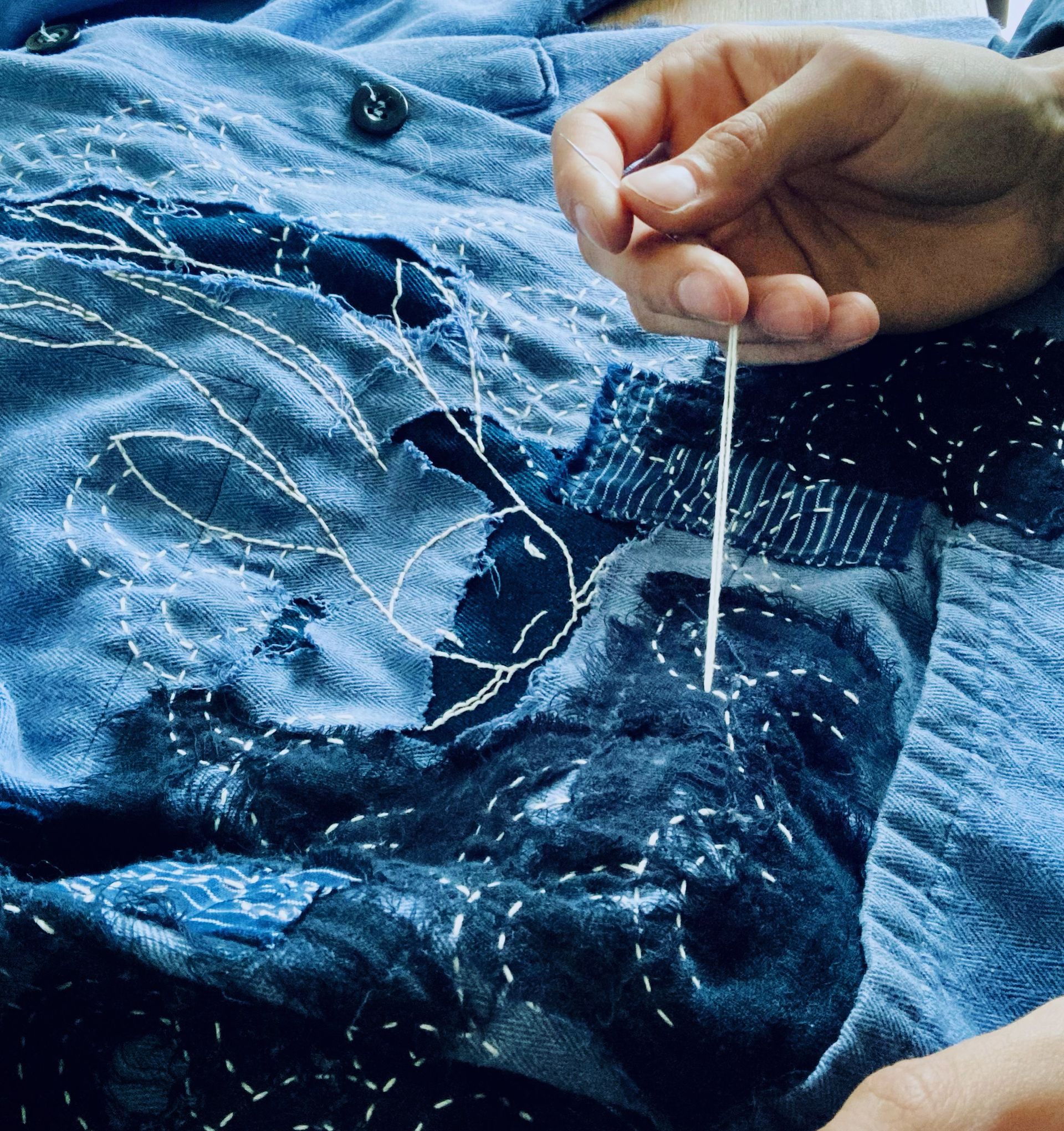 Hands creating white thread sashiko mending pattern on blue denim fabric by Sophie Cagniart