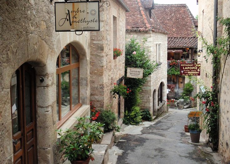 Narrow cobblestone alley with stone buildings, flower pots, and hanging shop signs in a quaint village.