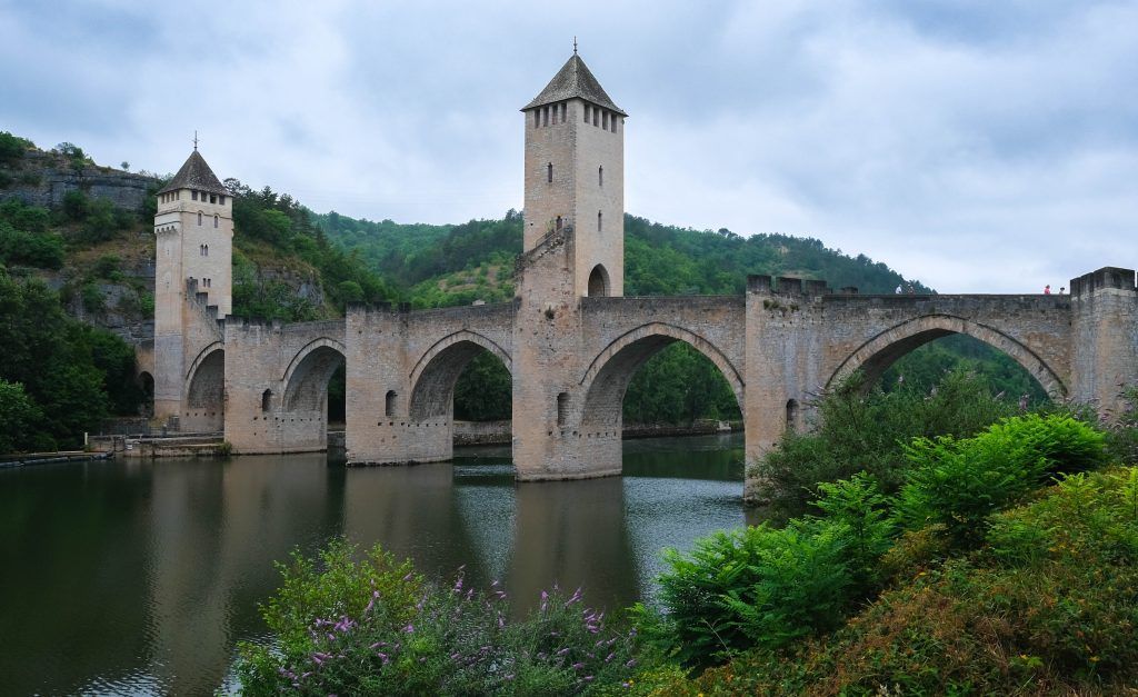 Stone bridge with towers over a river, surrounded by green hills under a cloudy sky