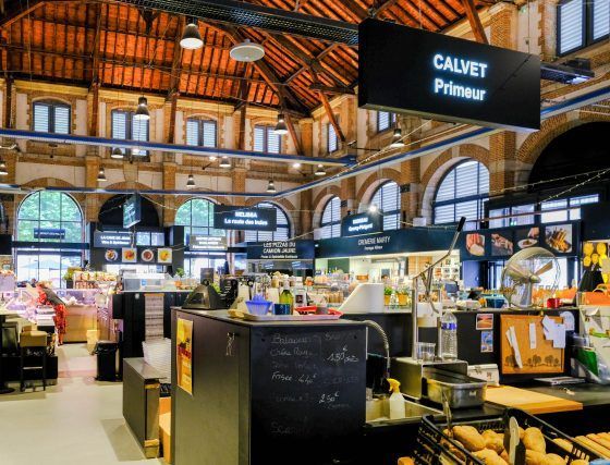Busy indoor market hall with arched brick ceiling and a Calvet Primur stall under digital signs