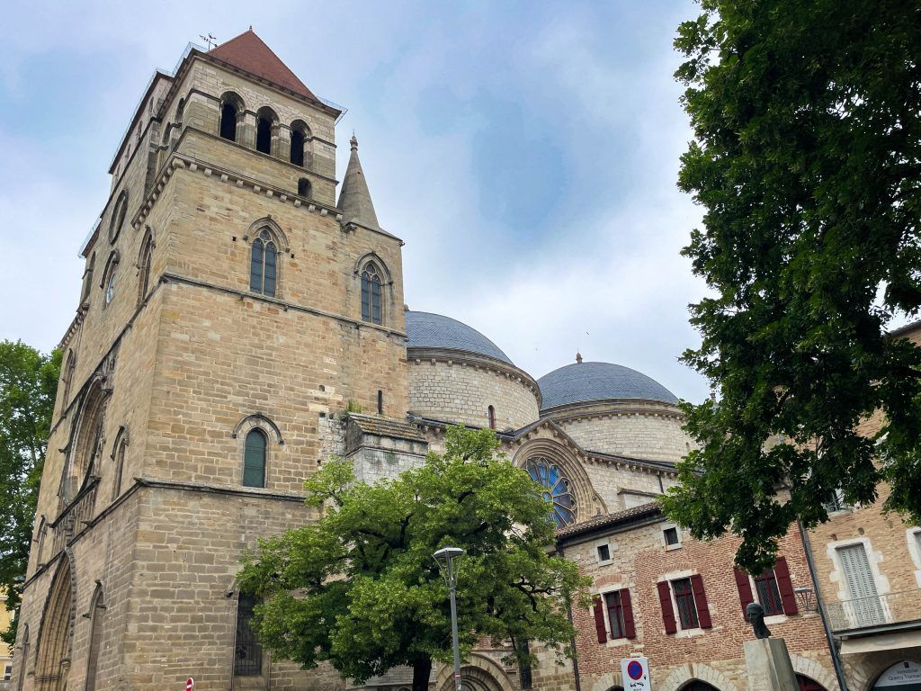 Historic stone church tower and domed roof behind trees and red-roofed buildings under a cloudy sky