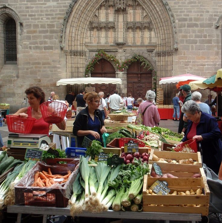 Outdoor French market stall with fresh vegetables in front of a stone building and shoppers browsing
