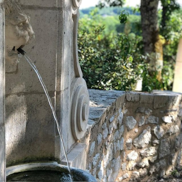 Stone fountain spout pouring water beside a sunlit garden wall and greenery