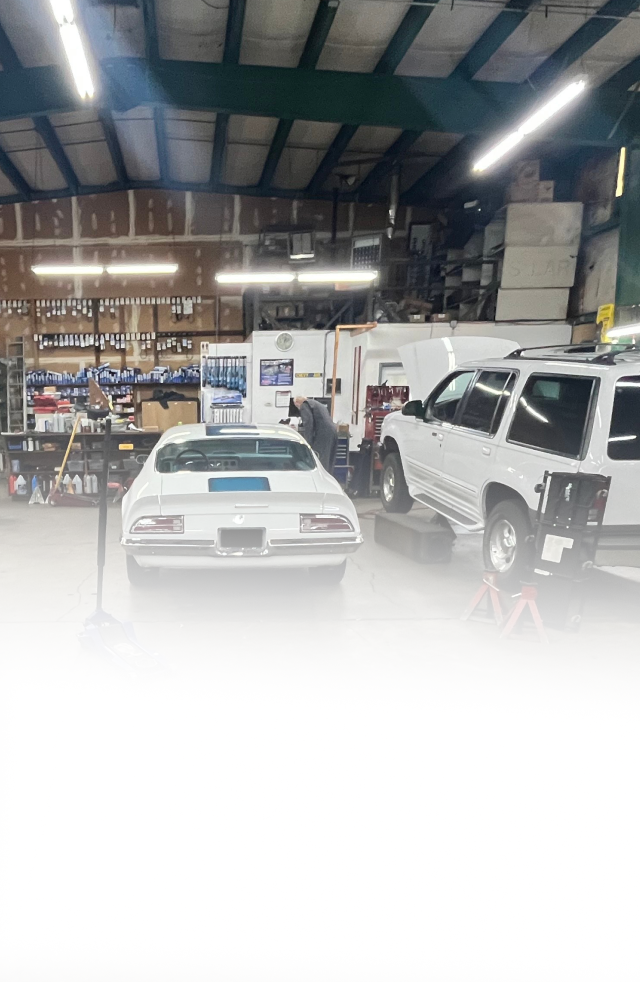 Two white vehicles parked in a large, well-lit automotive garage filled with parts and equipment.
