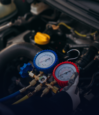 Gloved hands holding an AC manifold gauge set to check pressure levels on a car engine. | Maynard's Auto Repair Service