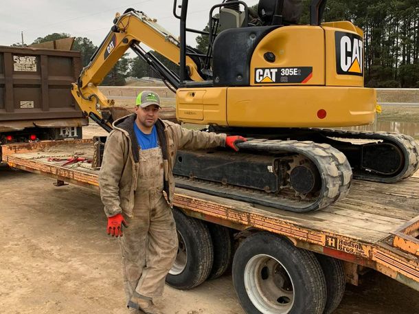 A man is standing next to a cat excavator on a trailer.