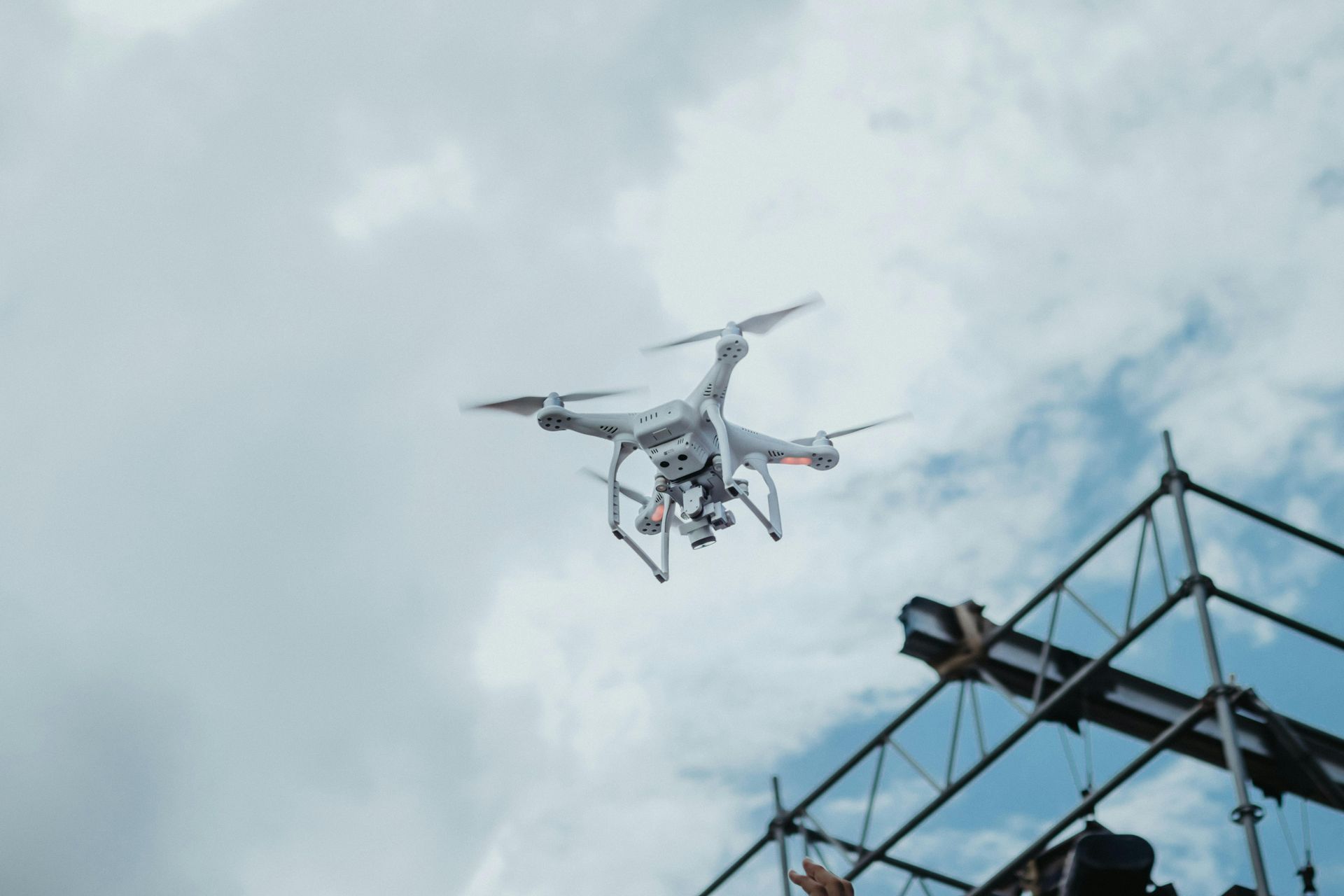 Drone flying in cloudy sky, near metal scaffolding.