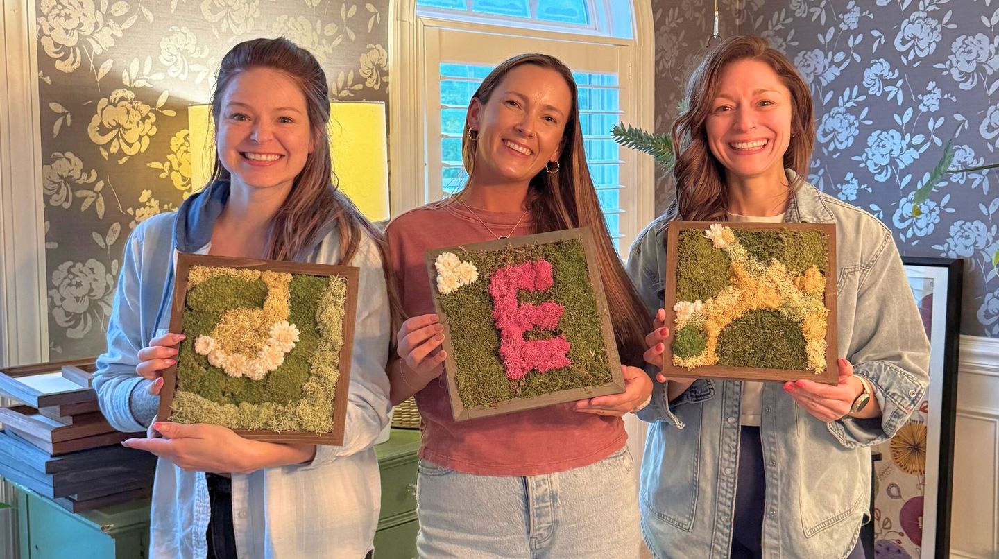 Three women smiling, holding framed floral art, indoors. One frame has an