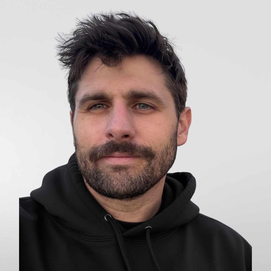 A person with short brown hair and light stubble wearing a tan baseball cap and black hoodie against a pale background.