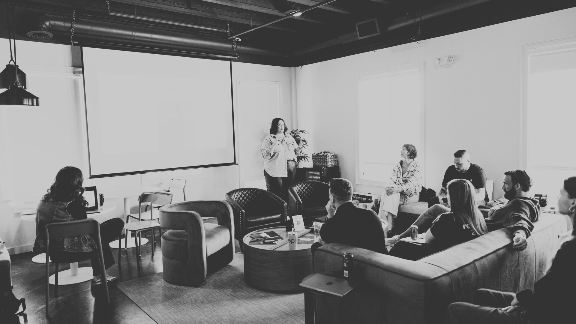 Group meeting in a modern office. Person presenting to seated colleagues, screen in background.