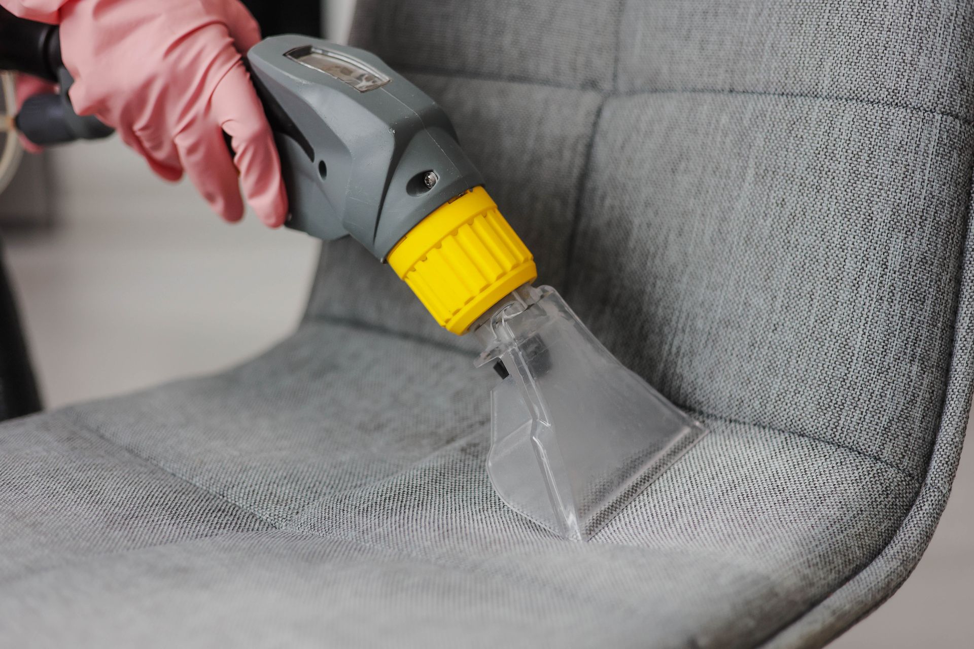 Person wearing pink gloves cleaning a gray upholstered chair with a handheld upholstery cleaner.