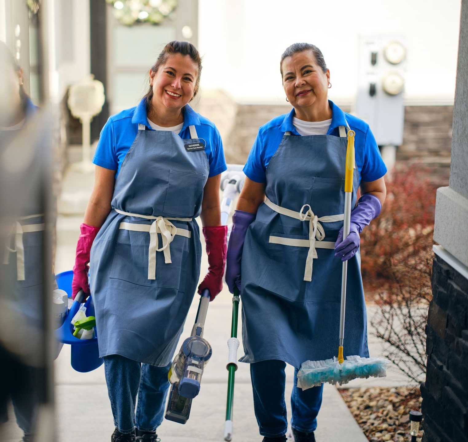 Two people wearing blue shirts and aprons holding cleaning supplies, smiling outside a building.