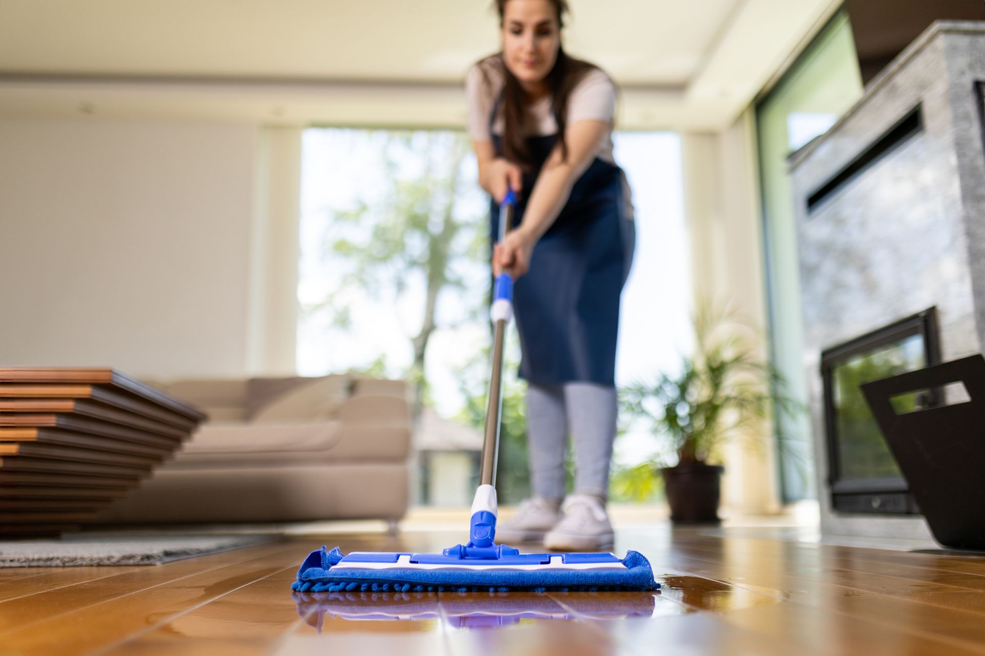 Woman mopping a wooden floor in a sunlit living room. Blue mop, gray leggings, and white top.