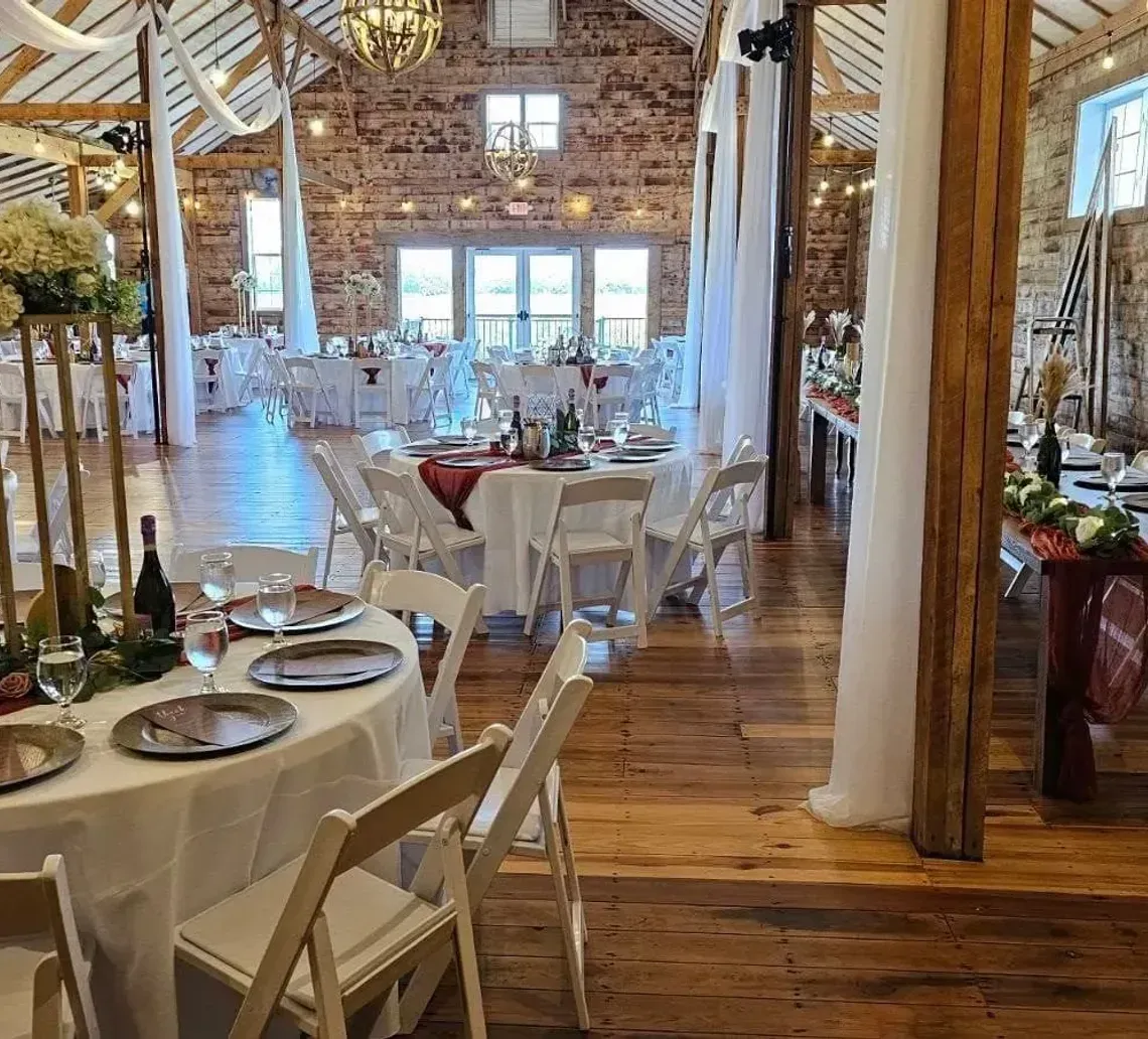 A rustic event venue featuring circular dining tables with white linens, floral centerpieces, and warm string lighting.