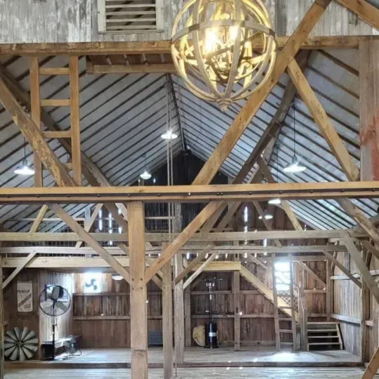 The rustic interior of a wooden barn features exposed rafters, a large sphere-shaped chandelier, and stairs to a loft.