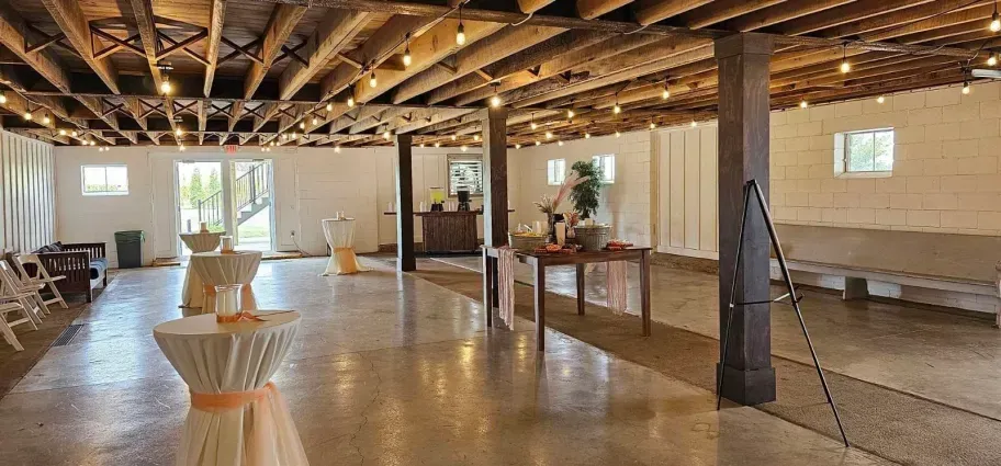 Rustic event space with white walls, exposed ceiling beams, string lights, a concrete floor, and scattered tables.